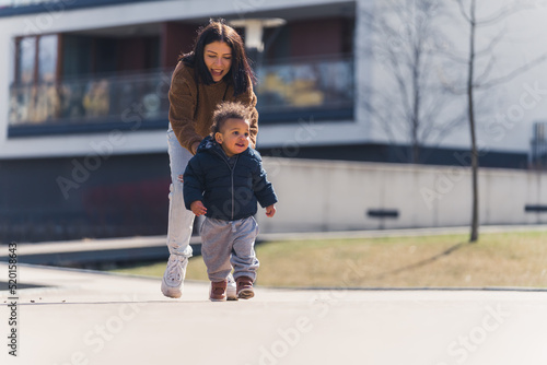 Young caucasian Single mother's cute african little baby running on the path of residential area, both wearing warm winter clothes - modern building background. High quality photo
