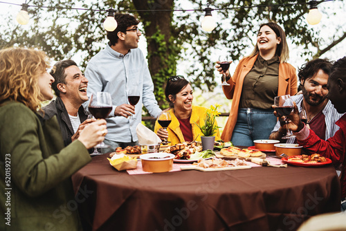 Group of multiethnic carefree friends talk, joke and drink wine during the brunch of the harvest wine tasting, sitting at the table on the terrace of the farmhouse vinery 