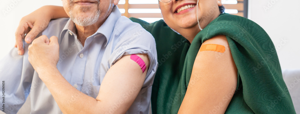 Showing bandage on arm, shoulder, happy asian elderly, aged family smile strong together with protect of covid-19, coronavirus after injection of vaccine, sitting on couch in living room at home.