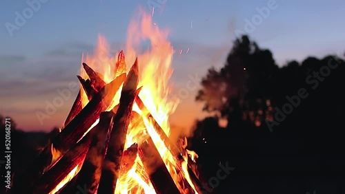 Big Burning Campfire at Summer Evening against the Blue Sky. Wood on Fire. Flying Sparks. Travel and Tourism Concept. Giant Flaming Bonfire - Static Shot, Slow Motion