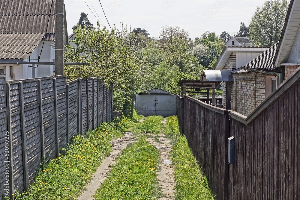 rural alley with a road in green grass along fences made of gray concrete and brown wooden boards and gray metal gates in a dead end rural alley with a road in green grass along fences made of gray concrete and brown wooden boards and gray metal gates in a dead end