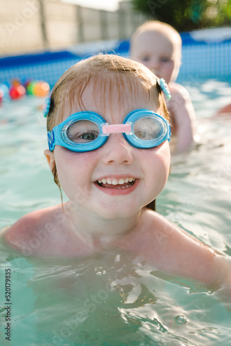 Little baby in swimming goggles, girl swim in paddling pool. Diving kid. Learning child to swim. Summer leisure and family holidays and vacation concept. People swim in a metal frame pool in backyard. © Serhii