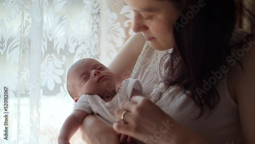 mother holding in her arms and caressing her newborn baby by the window