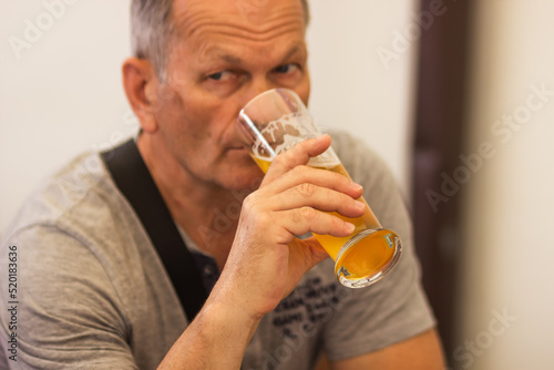 Caucasian senior man with gray hair in gray t-shirt holding and drinking glass of light beer in craft czech brewery.