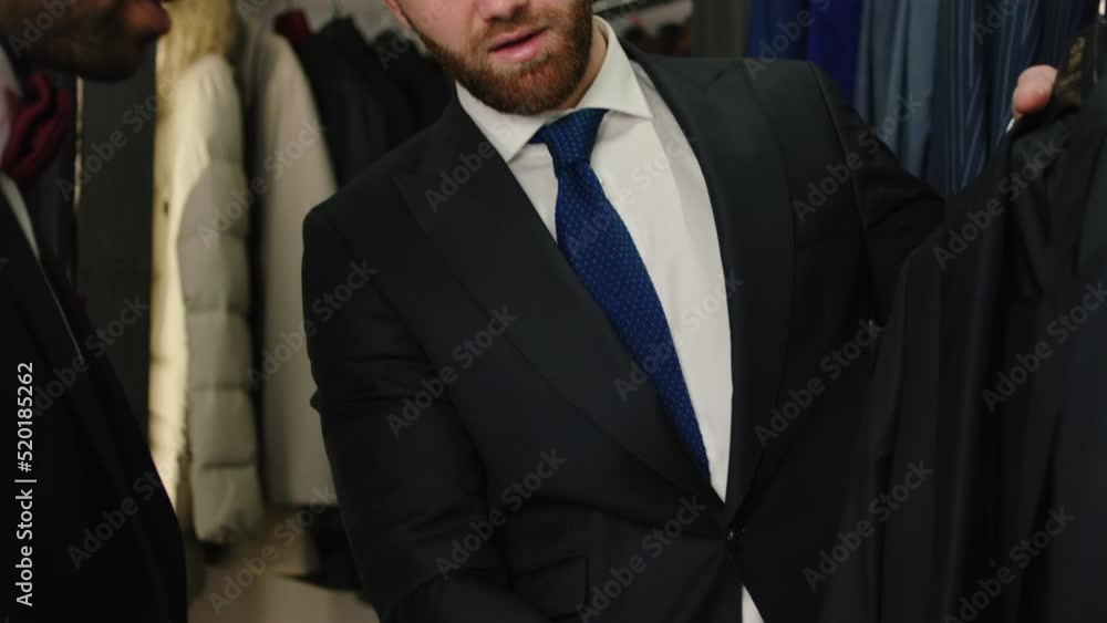 Handsome man groom try to choose the good and best suit for the wedding together with the shop consultant man they looking at the new suit from the collection