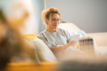 Young mature woman smile and enjoy time reading an electronic book on tablet reader device comfortably sitting on the sofa at home in indoor leisure activity alone. Female people have relax indoor