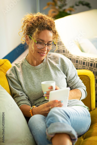 Woman at home have fun and enjoy reading e-book from modern reader. Female have leisure activity indoor in apartment flat laying on a yellow couch and drinking coffee alone with a smile. Lifestyle