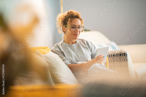 Young mature woman smile and enjoy time reading an electronic book on tablet reader device comfortably sitting on the sofa at home in indoor leisure activity alone. Female people have relax indoor photo