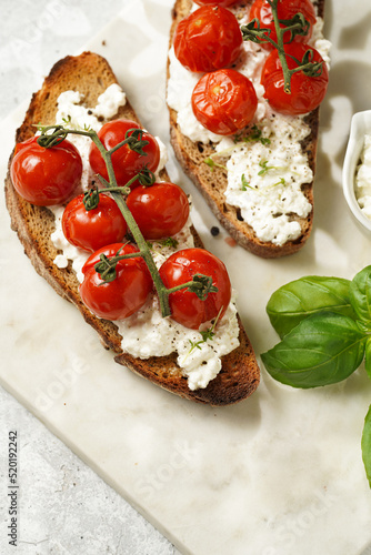 A sandwich with roasted cherry tomatoes with branch, fresh cottage cheese, green basil on a slice of whole wheat bread on a marble board on grey background