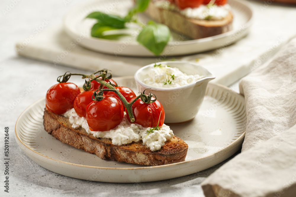 A sandwich with roasted cherry tomatoes with branch, fresh cottage cheese, green basil on a slice of whole wheat bread on a round plate on grey background