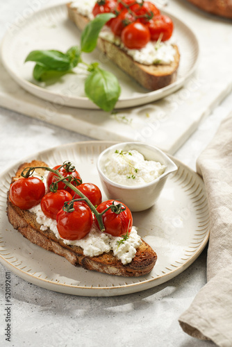 A sandwich with roasted cherry tomatoes with branch, fresh cottage cheese, green basil on a slice of whole wheat bread on a round plate on grey background