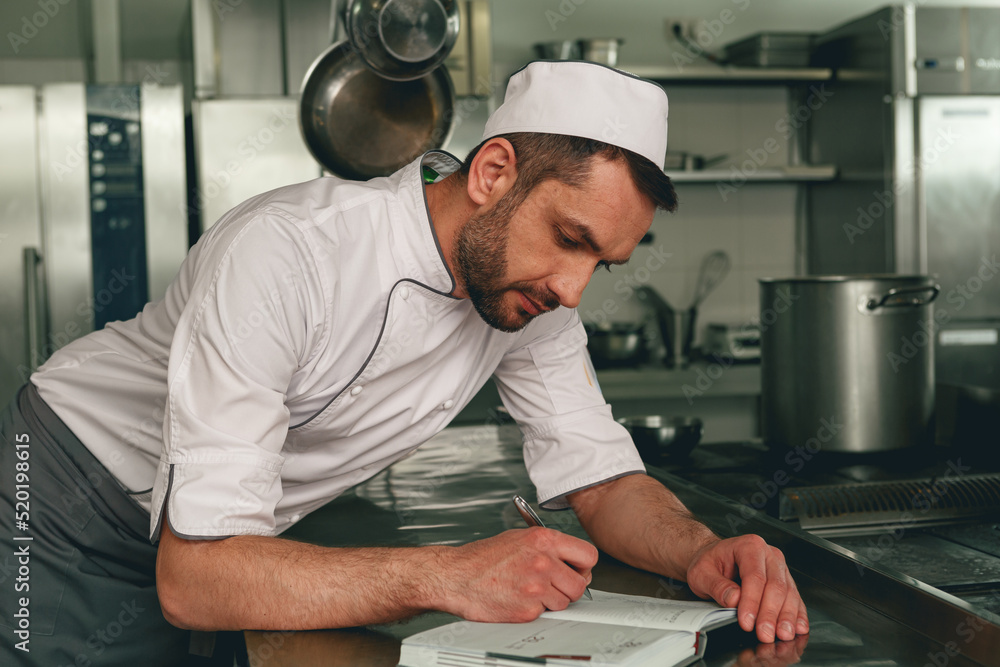 Smiling chef in uniform making notes in notebook standing on kitchen