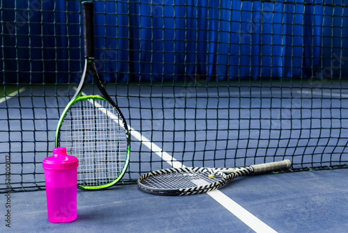 Tennis rackets and a water bottle against a stretched tennis net on an indoor tennis court © ribalka yuli