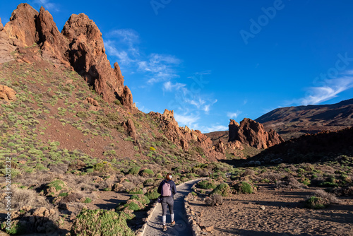 Woman with backpack on hiking trail with scenic golden hour sunrise morning view on unique rock formation at Roques de Garcia, Tenerife, Canary Island, Spain, Europe. Pico del Teide volcano summit