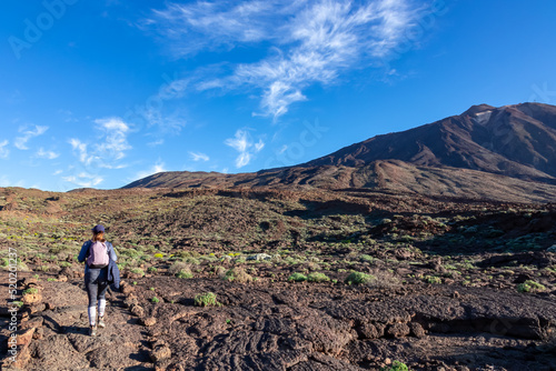 Woman with backpack on hiking trail with panoramic view on summit of Pico del Teide, Mount El Teide National Park, Tenerife, Canary Island, Spain, Europe. Walking over barren volcanic stone terrain