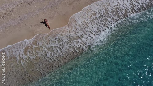 Aerial view of beautiful happy woman in swimsuit sitting on sand in the shallow sea water enjoying beach and soft turquoise ocean wave. Tropical sea in summer season on Egremni beach on Lefkada island