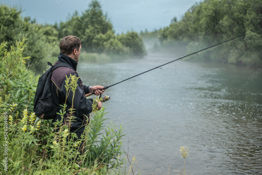Male fisherman is standing on the riverbank with a fishing rod in his hands and catching fish. Calm outdoor recreation Male fisherman is standing on the riverbank with a fishing rod in his hands and catching fish. Calm outdoor recreation