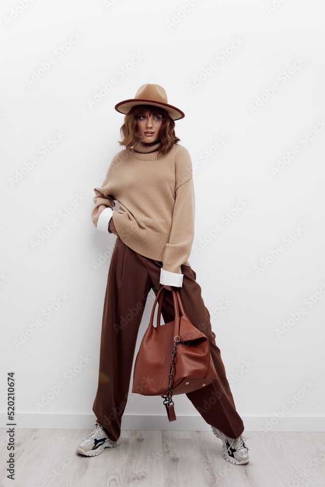 a beautiful woman stands full-length on a light background in a hat, sweater and shirt, legs wide apart, holding a bag in one hand, putting her hand on her belt