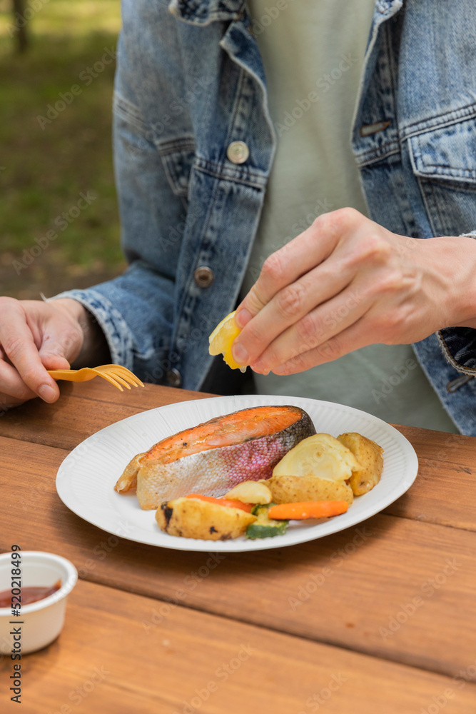 Salmon grilled fish with grilled vegetables in eco disposable tableware. Grilled potato, carrot, vegetable marrow. Man 
water salmon with lemon. Picnic in park. Vertical photo. 