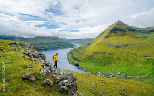 Male photographer stands on the edge of a cliff overlooking the Funningur fjord on Faroe Islands. Stunning view of the magnificent green cliffs and mountains in the picturesque Faroe Islands.