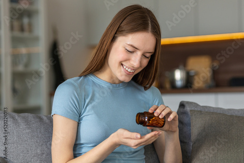 Happy pretty woman taking a pill sitting at home on the couch. Caucasian young female with bottle of pills, takes vitamins