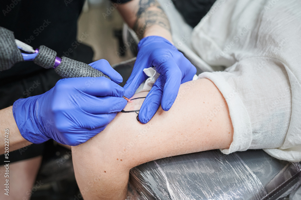 Fototapeta premium Unrecognizable tattooist starts a new tattoo design applying ink under the skin using a needle tattoo machine at the studio, wearing blue gloves.
