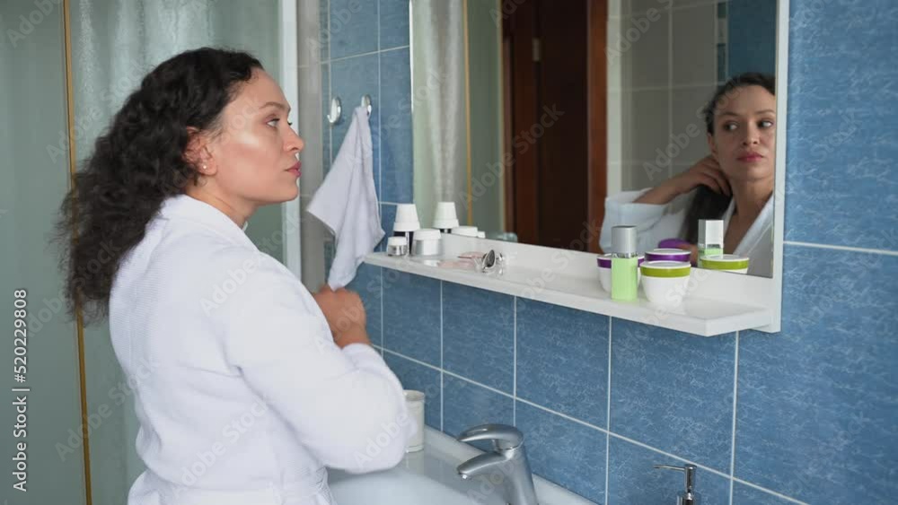 Dark haired mature woman, in a white waffle bathrobe, combing hair, looking at her reflection in the bathroom mirror Dark haired mature woman, in a white waffle bathrobe, combing hair, looking at her reflection in the bathroom mirror