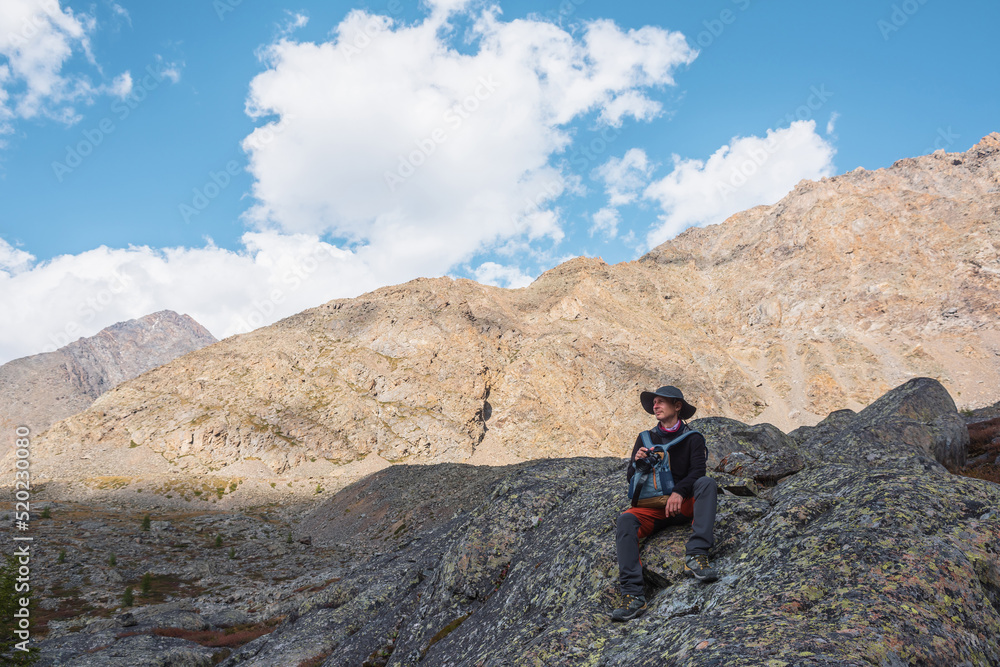 Hiker with backpack sitting on stone against rocks in bright sunlight. Alone man with photo camera in bright sun near sunlit rocky mountain wall. Tourist on moraines in high mountains in sunny day.