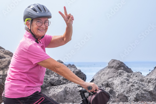 Active happy senior woman with helmet running on her electric bicycle along the sea looking at camera with victory sign. Freedom and vacations concept photo