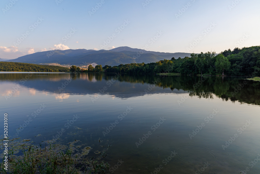 A lake reflects sunlight, against blue sky and mountain ranges and villages