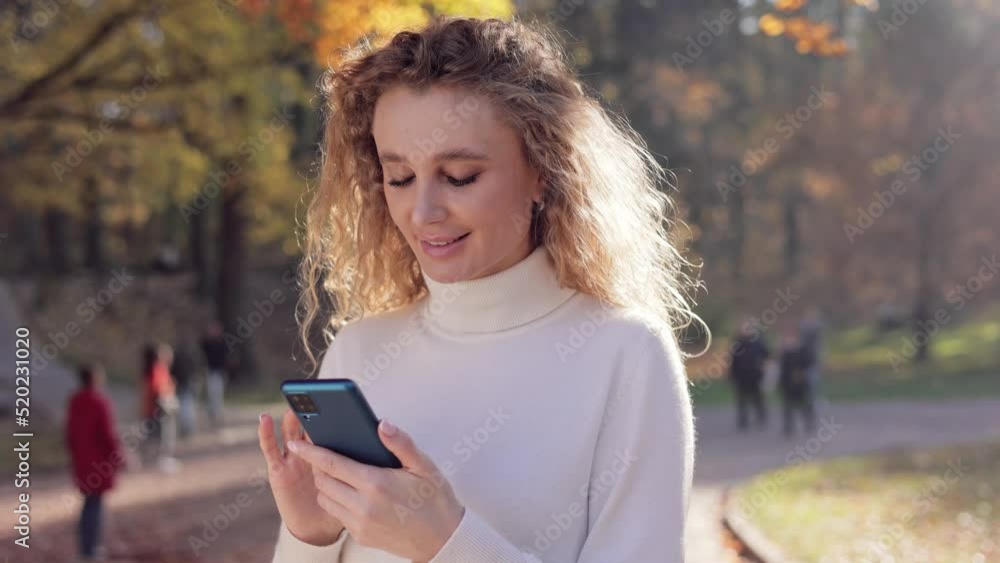 Portrait of beautiful blondie woman using smartphone in autumn park. Smiling young curly woman texting message, reading news, watching photo or video in social media walking at sunset in the park. Portrait of beautiful blondie woman using smartphone in autumn park. Smiling young curly woman texting message, reading news, watching photo or video in social media walking at sunset in the park.