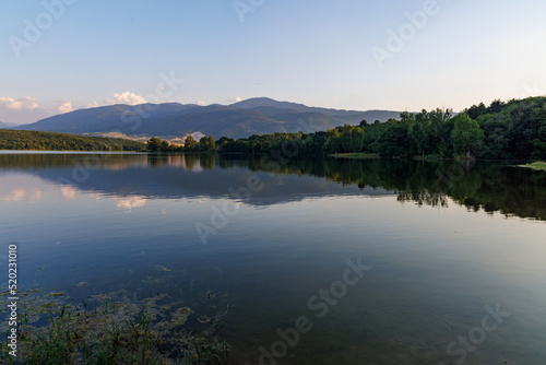 A lake reflects sunlight, against blue sky and mountain ranges and villages