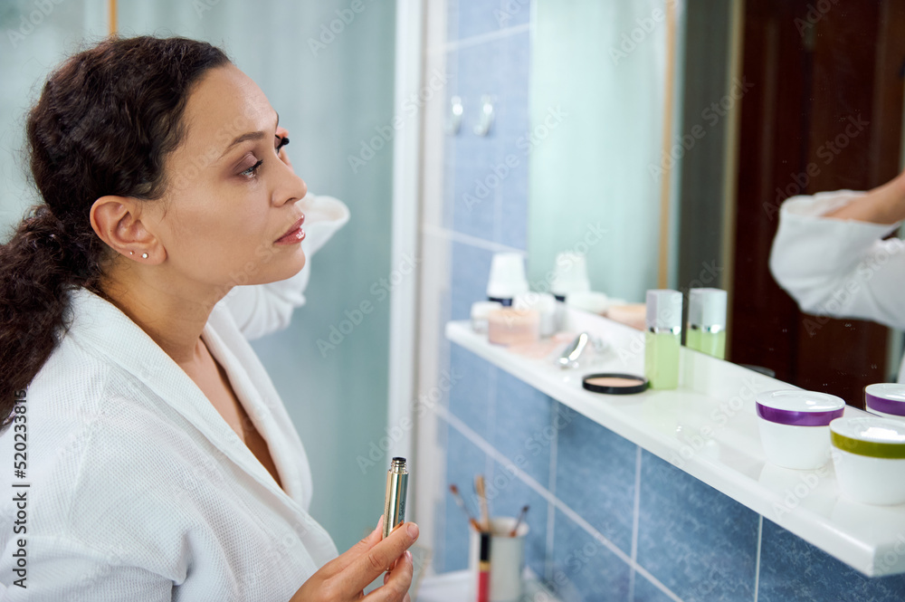 Fototapeta premium Charming multiethnic woman wearing a white bathrobe, applying make-up, using mascara, in front of a bathroom mirror