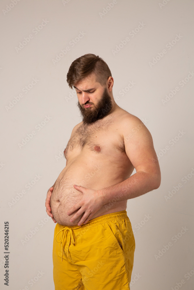Vertical photo with a pregnant man. A young man with a beard looks sadly at his fat belly. The bearded man holds his stomach with his hands. Fat man in yellow shorts on a gray background