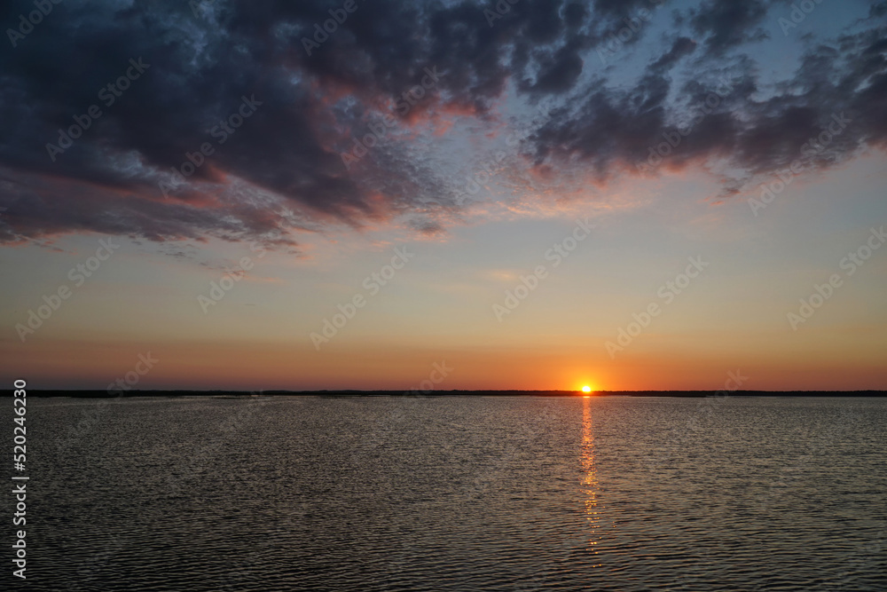 Sunset on the Łebsko lake, Pomeranian Voivodeship, Poland. Summer evening, dramatic dark clouds in the sky, the sun hides behind the horizon, the reflection of light in the water. Sunset on the Łebsko lake, Pomeranian Voivodeship, Poland. Summer evening, dramatic dark clouds in the sky, the sun hides behind the horizon, the reflection of light in the water.