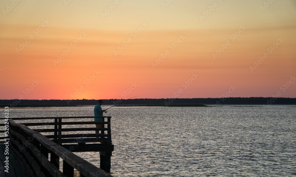 Angler silhouette on the pier at sunset in summer at Lebsko Lake, Pomerania, Poland, Europe.