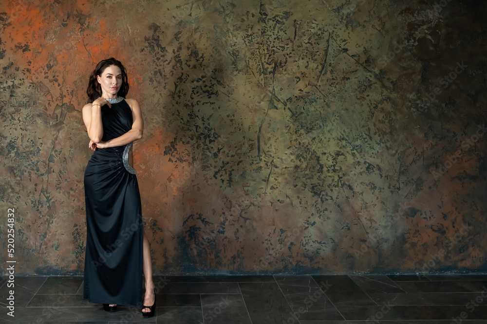 Beautiful caucasian brunette-haired woman wearing black evening gown with a slip stands by dark wall. Copy space for your text. People theme.