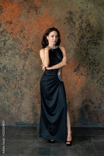 Beautiful caucasian brunette-haired woman wearing black evening gown with a slip stands by dark wall. Beautiful people theme.