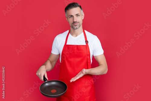 mature man chef in apron with frying pan on red background. presenting product photo