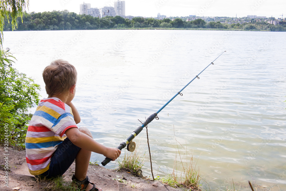 child is fishing in river on sunny summer day. young fisherman boy cast a fishing rod and is sitting waiting for a bite, view of river. Nature. Outdoor. Happy and interesting childhood child is fishing in river on sunny summer day. young fisherman boy cast a fishing rod and is sitting waiting for a bite, view of river. Nature. Outdoor. Happy and interesting childhood