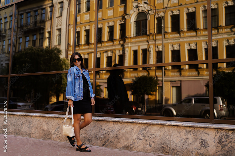 Side view of stylish woman a black dress and jeans jacket strolling along street near a vintage building . 
Modern lady in sunglasses walks in city . Street style shooting. Women's fashion . 