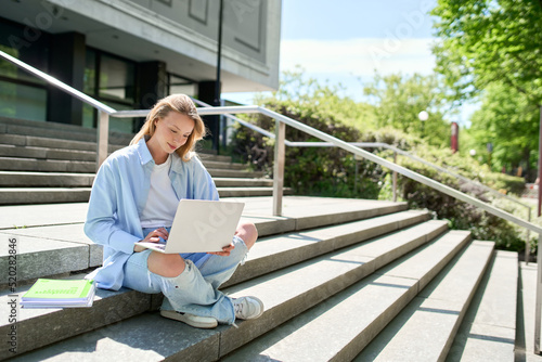 Fotografie Smiling girl student using laptop computer modern technology device outdoor in university campus online learning, elearning outside sitting on urban stairs