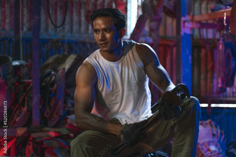 Asian mechanic sits in a red-blue rear light spare room in the garage. Asian mechanic sits in a red-blue rear light spare room in the garage.