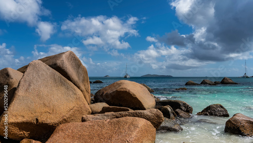 Huge picturesque boulders on the coast of the Indian Ocean are washed by turquoise water. Ships on the horizon. Clouds in the blue sky. Seychelles. Praslin Island. Anse Lazio beach