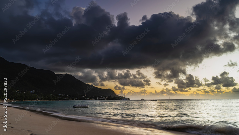 Sunset over the Indian Ocean. There are purple clouds in the orange-lit sky. Foam of waves on a sandy beach. The silhouette of a mountain in the distance. Seychelles. Mahe Island. Beau Vallon