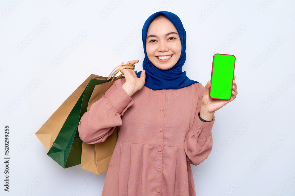 Cheerful young Asian muslim woman in pink shirt holding package bags with purchases after shopping and showing blank screen mobile phone isolated on white background. People lifestyle concept Cheerful young Asian muslim woman in pink shirt holding package bags with purchases after shopping and showing blank screen mobile phone isolated on white background. People lifestyle concept