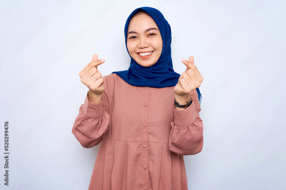 Smiling young Asian muslim woman in pink shirt showing Korean heart with two fingers crossed, express joy and positivity isolated over white background. People religious lifestyle concept