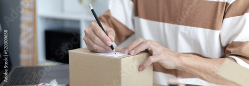 Young Asian man is writing down the customer's details and addresses on the notebook or box in order to prepare for shipping according to the information, packing box, Sell online, freelance working.