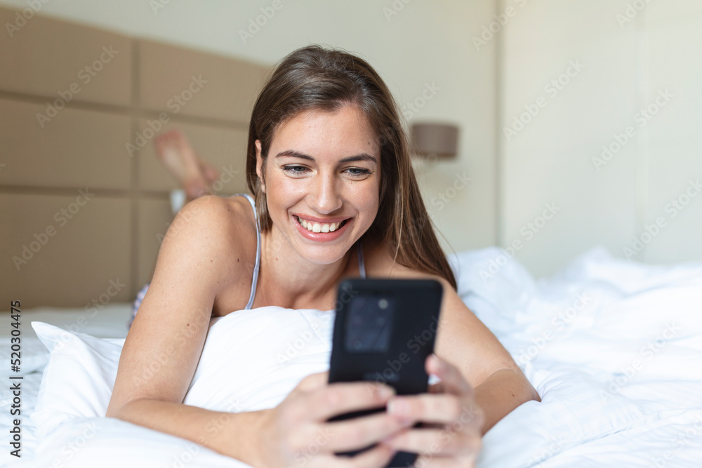 Smiling woman looking at mobile phone while laying on white bed. Happy brunette young woman using cellphone at home. Beautiful girl surfing the net on smartphone Smiling woman looking at mobile phone while laying on white bed. Happy brunette young woman using cellphone at home. Beautiful girl surfing the net on smartphone