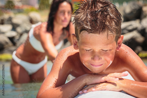 Cheerful teenage boy lying on a swimming board. Happy european family having fun floating on a swim board in the sea on a hot summer day
